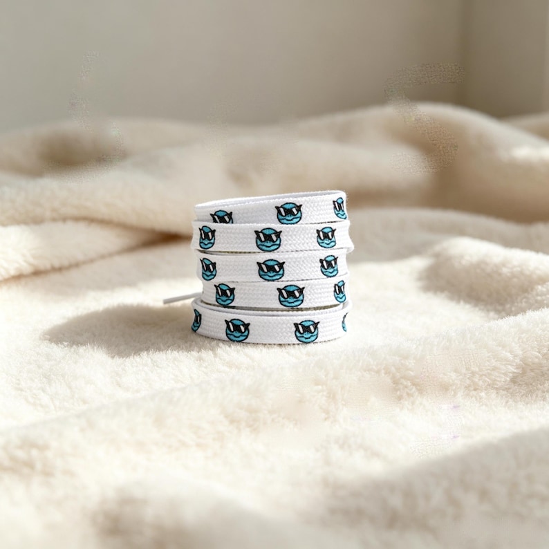 May include: A white shoelace coiled into a stack, featuring a repeating pattern of small, light blue circular faces wearing black sunglasses. The shoelace is flat and woven, with the graphic print evenly spaced along the entire length of the fabric.