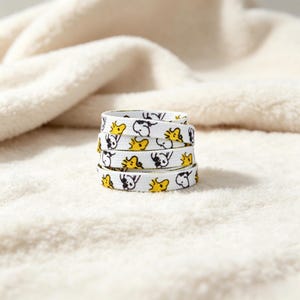 May include: A coiled white shoelace featuring a repeating pattern of small black and white dog illustrations and yellow bird illustrations. The flat fabric lace is wound into a circular stack, displaying the graphic print across its entire surface.