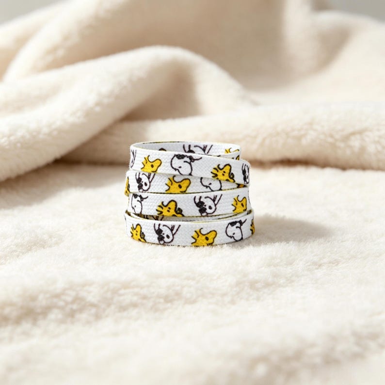 May include: A coiled white shoelace featuring a repeating pattern of small black and white dog illustrations and yellow bird illustrations. The flat fabric lace is wound into a circular stack, displaying the graphic print across its entire surface.