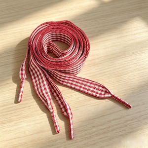 Può includere: Un paio di lacci piatti con un motivo a quadretti vichy rosso e bianco. I lacci sono arrotolati in forma circolare, con puntali in plastica trasparente alle estremità. Il tessuto ha una trama intrecciata e il motivo è costante.