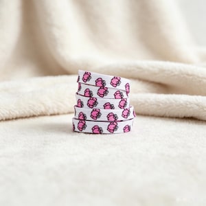 May include: A coiled white shoelace featuring a repeating pattern of small pink crabs with black eyes. The flat fabric lace is wound into a stack, displaying the consistent graphic print across the visible segments of the material.