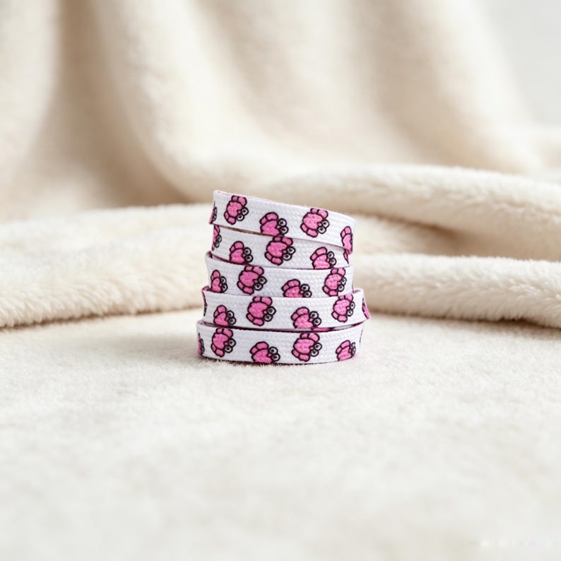 May include: A coiled white shoelace featuring a repeating pattern of small pink crabs with black eyes. The flat fabric lace is wound into a stack, displaying the consistent graphic print across the visible segments of the material.