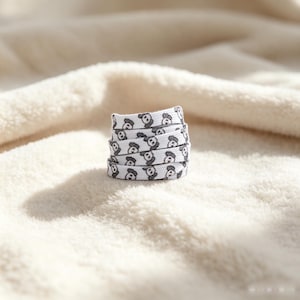 May include: A white flat shoelace coiled into a small stack, featuring a repeating pattern of small, dark gray cartoon bear faces with X-shaped eyes. The shoelace is made of a woven fabric material and displays a consistent graphic print throughout.