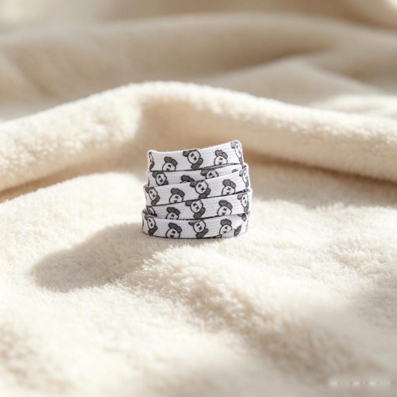 May include: A white flat shoelace coiled into a small stack, featuring a repeating pattern of small, dark gray cartoon bear faces with X-shaped eyes. The shoelace is made of a woven fabric material and displays a consistent graphic print throughout.