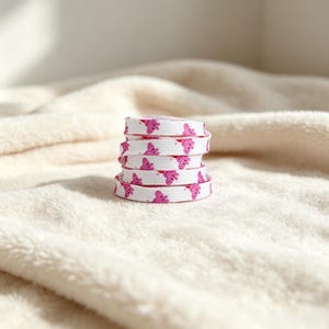 May include: A coiled white shoelace featuring a repeating pattern of small pink cartoon pig characters. The shoelace is woven with a flat texture and displays the pink pig graphics clearly across the white fabric surface in a stacked circular form.