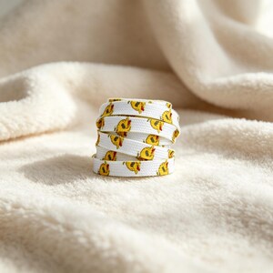 May include: A white shoelace coiled into a cylindrical stack, featuring a repeating pattern of small yellow cartoon ducklings with orange beaks. The shoelace has a flat, woven texture and is displayed as a single, neatly rolled unit.