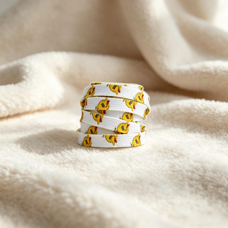 May include: A white shoelace coiled into a cylindrical stack, featuring a repeating pattern of small yellow cartoon ducklings with orange beaks. The shoelace has a flat, woven texture and is displayed as a single, neatly rolled unit.