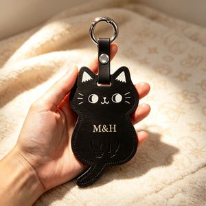 May include: A black cat-shaped leather keyring featuring white printed facial details, including eyes, whiskers, and ears. The centre of the cat body displays the letters M&H in a gold-coloured font. A silver metal ring is attached to the top.