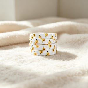 May include: A coiled white shoelace featuring a repeating pattern of small yellow and brown cartoon character heads. The shoelace is made of a flat woven fabric and is arranged in a circular stack, displaying the graphic print across its surface.