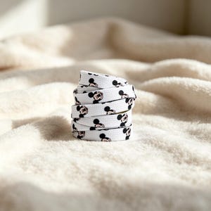 May include: A coiled white shoelace featuring a repeating pattern of black and pink Mickey Mouse character heads. The shoelace is made of a flat woven fabric material and is arranged in a vertical stack, displaying the graphic print clearly.