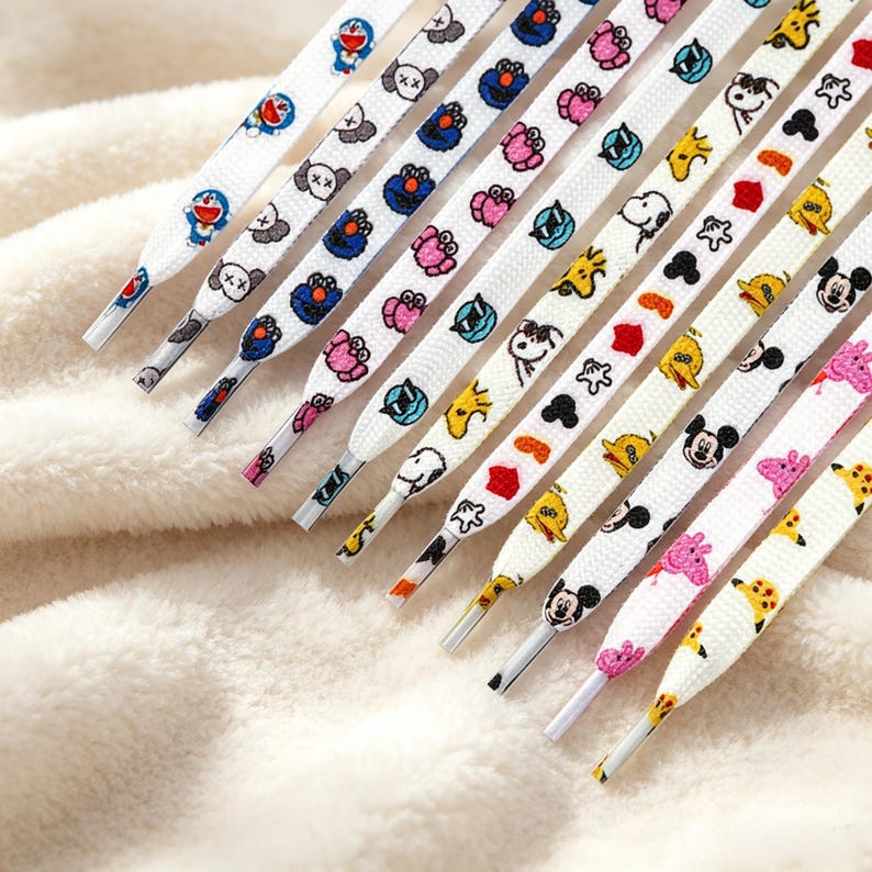 May include: A collection of white shoelaces featuring various colorful cartoon character prints, including Doraemon, Cookie Monster, Snoopy, Mickey Mouse, and Peppa Pig. Each lace has a smooth, flat woven texture with clear plastic aglets at the ends.