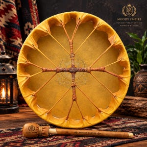May include: A round yellow drum with a tan drumhead featuring a central cross-shaped lacing pattern made of thin leather strips. A matching wooden drum mallet with a rounded head and the text Moody Empire is positioned horizontally in front.