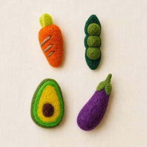May include: Four felted vegetable shapes including an orange carrot with green top, a dark green pea pod with three round peas, a halved avocado with a yellow center and brown pit, and a purple eggplant with a green stem on a textured fabric.