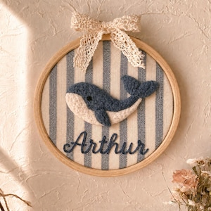 Peut inclure: Un cadre circulaire en bois pour broderie contenant un tissu à rayures verticales bleues et blanches. Une baleine en feutrine bleu foncé et crème est centrée, avec le nom Arthur brodé en bleu foncé en dessous et un nœud en dentelle.