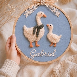 Op de afbeelding: Een ronde houten borduurring met blauwe stof en een naaldviltontwerp van een grote gans met een donkergroene sjaal en een klein gansje. De naam Gabriel is in witte schrijfletters eronder geborduurd, afgewerkt met een kanten strik.