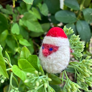 Pode incluir: Uma figura de Pai Natal em lã feltrada com um gorro vermelho, barba branca e pequenos olhos azuis. O ornamento redondo e texturizado apresenta um nariz vermelho proeminente e é feito com um acabamento macio e felpudo.