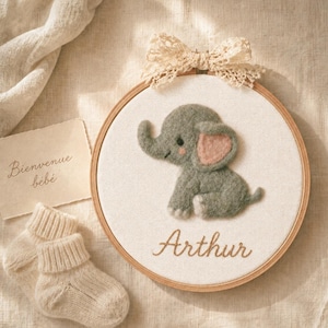 May include: A circular wooden embroidery hoop containing a needle-felted gray elephant with pink ears on white fabric. The name Arthur is embroidered in brown script below the elephant. A cream lace bow is attached to the top of the hoop.