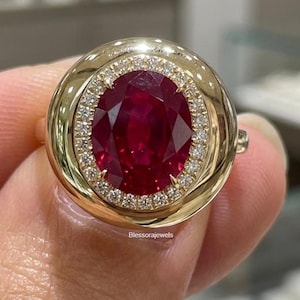 Peut inclure: Une bague en or avec un grand rubis rouge foncé taille ovale au centre, entouré d'un halo de petits diamants ronds transparents. La monture est un anneau en or poli et lisse avec un profil large et arrondi. Le texte Blessorajewels est visible.