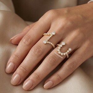 Puede incluir: Un anillo de oro con un diseño abierto de dos secciones distintas, cada una adornada con una serie de pequeñas piedras redondas y transparentes en engaste de bisel. El texto Blessorajewels es visible en la parte inferior del anillo de oro.
