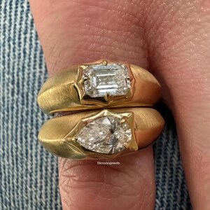 Peut inclure: Deux bagues en or avec une finition mate brossée, chacune dotée d'une pierre précieuse transparente sertie dans un chaton. La bague supérieure porte une pierre rectangulaire taille émeraude, et la bague inférieure une pierre en forme de poire. Texte : Blessorajewels.
