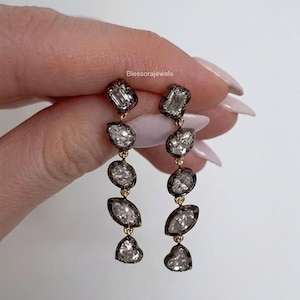 May include: A pair of drop earrings featuring five faceted clear stones in dark metal settings, arranged vertically in varying shapes including rectangular, pear, oval, marquise, and heart, connected by small gold links with the text Blessorajewels.