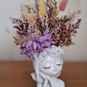 Peut inclure: Un cache-pot en céramique blanche en forme de tête représentant un personnage aux yeux fermés, les mains posées sous le menton. Il est garni d'un arrangement de fleurs séchées comprenant des hortensias violets, des queues-de-lapin et des herbes.