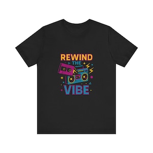 May include: A black short-sleeve t-shirt featuring a graphic print of a blue boombox and a pink cassette tape. The text REWIND THE VIBE is printed in yellow and blue letters above and below the graphic, surrounded by small geometric shapes.