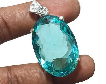 May include: An oval-shaped, faceted, translucent teal gemstone pendant set in a silver-toned metal prong setting. The stone features a brilliant cut with multiple reflective surfaces, and the top includes a decorative silver-toned bail.