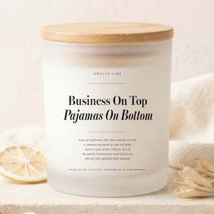 Peut inclure: Une bougie en verre dépoli avec un couvercle en bois clair. L'étiquette blanche porte l'inscription "SMELLS LIKE Business On Top Pajamas On Bottom" suivie d'un texte descriptif sur les appels en conférence, les trajets et le télétravail.