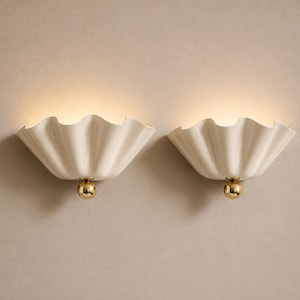 May include: A pair of wall lights featuring off-white, ruffled, shell-shaped shades. Each light is accented with a polished gold-tone spherical detail at the base, emitting a soft, warm glow from the top of the curved, pleated ceramic structure.