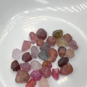 Puede incluir: Una colección de pequeñas piedras preciosas en bruto y sin pulir en varios tonos de rosa, rojo, morado y marrón. Estos especímenes minerales irregulares, de translúcidos a opacos, están dispersos y muestran facetas naturales.