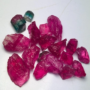 May include: A collection of raw, unpolished gemstones featuring numerous vibrant magenta-pink crystals alongside two smaller, distinct pieces that transition from a deep green base to a translucent, light-colored top section.