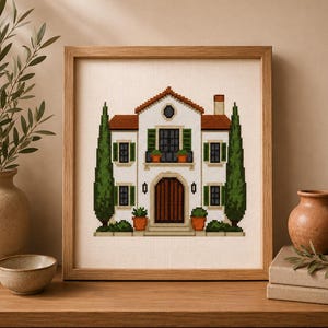 May include: A cross-stitch embroidery in a wooden frame depicting a white two-story house with a brown roof, a central arched wooden door, green shutters, and two tall green cypress trees flanking the structure, set against a light fabric background.