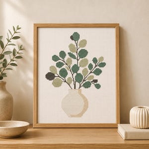 May include: A framed cross-stitch embroidery featuring a plant with round leaves in shades of dark green, sage, and olive, arranged in a light beige vase. The design is set against a white grid background and enclosed in a light wood frame.