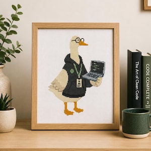 Może przedstawiać: Oprawiony haft krzyżykowy przedstawiający gęś w stylu pixel art, noszącą okrągłe okulary, czarną bluzę z kapturem i zieloną smycz z identyfikatorem. Gęś trzyma mały szary laptop na tle białego tła.