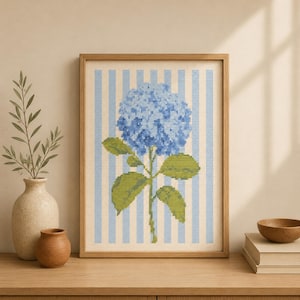 以下が含まれることがあります： 縦のライトブルーと白のストライプの背景の中央に、大きな青いアジサイの花と緑の葉が描かれたクロスステッチ刺繍の額縁入りアート。作品はシンプルな明るい色調の木製フレームに収められています。