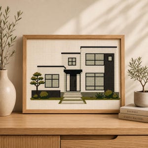 May include: A cross-stitch embroidery in a light wood frame depicting a modern two-story house with white walls, dark gray accents, large rectangular windows, a central front door, a set of stairs, and green shrubbery on a cream background.