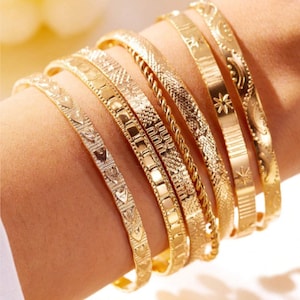 Puede incluir: Un conjunto de seis brazaletes de metal en tono dorado con varios patrones en relieve, incluyendo corazones, formas geométricas y estrellas. Las pulseras están apiladas, cada una mostrando una superficie texturizada y un acabado pulido.