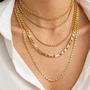 Puede incluir: Un conjunto de collares dorados en capas que presenta cinco cadenas distintas de varios estilos, incluyendo eslabones tipo cable, de bola y planos. El texto Lunivra JEWELRY shine in your own way es visible a la izquierda sobre un fondo claro.