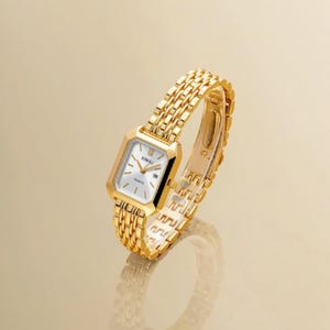 Puede incluir: Un reloj de pulsera dorado con caja rectangular y esfera blanca con índices dorados. El reloj tiene una correa de metal de eslabones múltiples. El texto Lunivra Jewelry shine in your own way es visible a la izquierda.