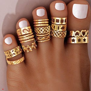 Puede incluir: Una colección de anillos para los dedos del pie en metal dorado, con varios diseños que incluyen corazones recortados, estrellas, patrones trenzados y formas geométricas. El texto Lunivra Jewelry shine in your own way es visible a la izquierda.