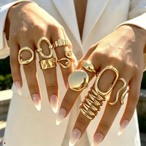 Könnte beinhalten: Eine Sammlung goldfarbener Ringe an den Fingern mit verschiedenen Formen, darunter offene Ovale, glatte Bänder, strukturierte Oberflächen, eine große runde Scheibe und ein mehrlagiges Spiraldesign. Der Text Lunivra Jewelry shine in your own way ist sichtbar.