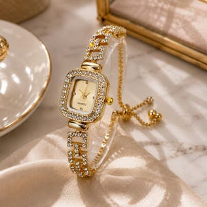 Puede incluir: Un reloj de cuarzo en tono dorado con esfera cuadrada y bisel incrustado con cristales transparentes. El reloj cuenta con una pulsera de eslabones a juego adornada con cristales y un mecanismo de cierre deslizante ajustable para la muñeca.