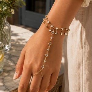 Könnte beinhalten: Ein goldfarbenes Handkettchen mit einer feinen Kette, die mit kleinen, klaren, rund geschliffenen Kristallen besetzt ist. Das Design umfasst ein Armband mit baumelnden Kristallakzenten und eine Verbindungskette, die zu einem Fingerring führt.