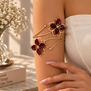 May include: A gold-toned arm cuff featuring two open-work flower motifs with deep burgundy petals and a central gold bead. The band has a textured finish and tapers to small spherical ends, wrapping around the upper arm in a spiral design.