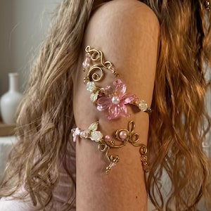 May include: Two gold-toned wire arm cuffs featuring pink floral accents, small white beads, and delicate leaf charms. The upper cuff showcases a large translucent pink flower with a central pearl, while the lower cuff displays smaller floral details.