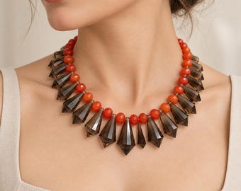 May include: A statement necklace featuring a strand of round, vibrant orange beads interspersed with faceted, dark gray, teardrop-shaped pendants. The design alternates between the bright orange spheres and the elongated, translucent gray crystals.