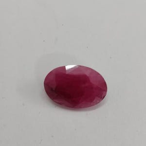 Può includere: Una gemma sciolta di forma ovale con un colore rosso profondo e saturo. La pietra presenta un taglio sfaccettato che crea molteplici superfici riflettenti sulla parte superiore, mostrando una qualità traslucida e un aspetto levigato.