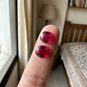 May include: Two oval-cut, deep red gemstones with faceted surfaces. The stones are translucent and display a vibrant crimson hue, reflecting light across their polished, multi-faceted tops. Each gem features a symmetrical oval shape.