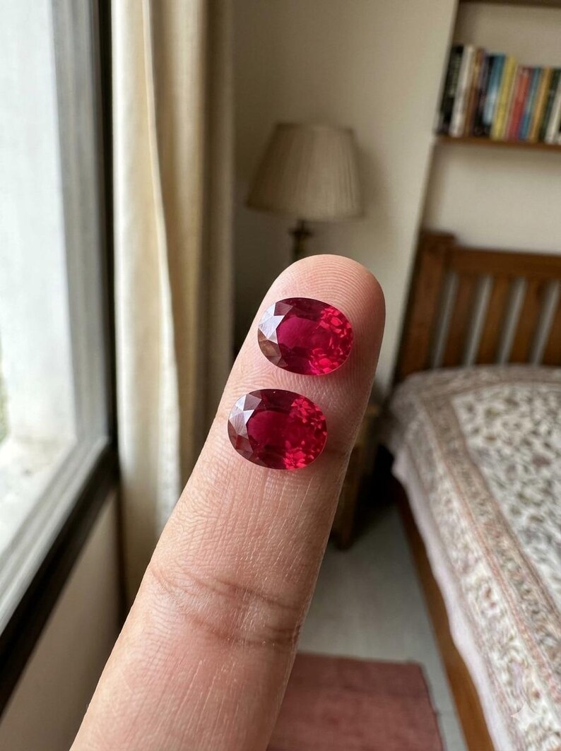 May include: Two oval-cut, deep red gemstones with faceted surfaces. The stones are translucent and display a vibrant crimson hue, reflecting light across their polished, multi-faceted tops. Each gem features a symmetrical oval shape.